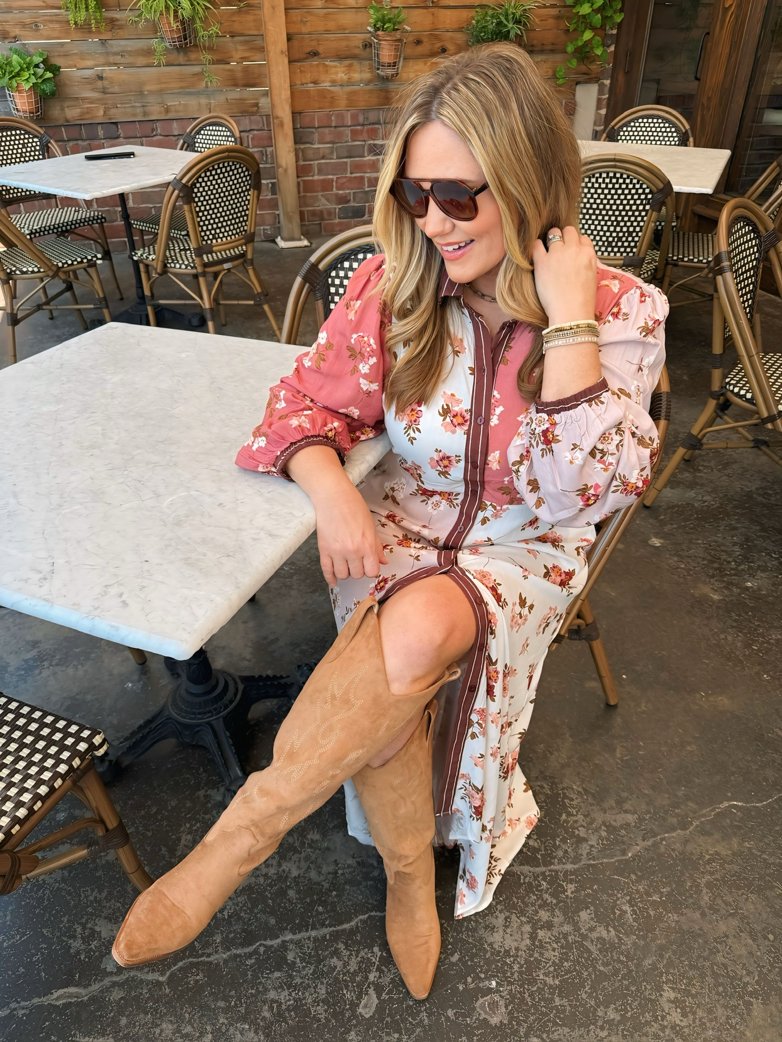 Audrie Dollins in floral dress and tan suede boots sitting at outdoor café table.