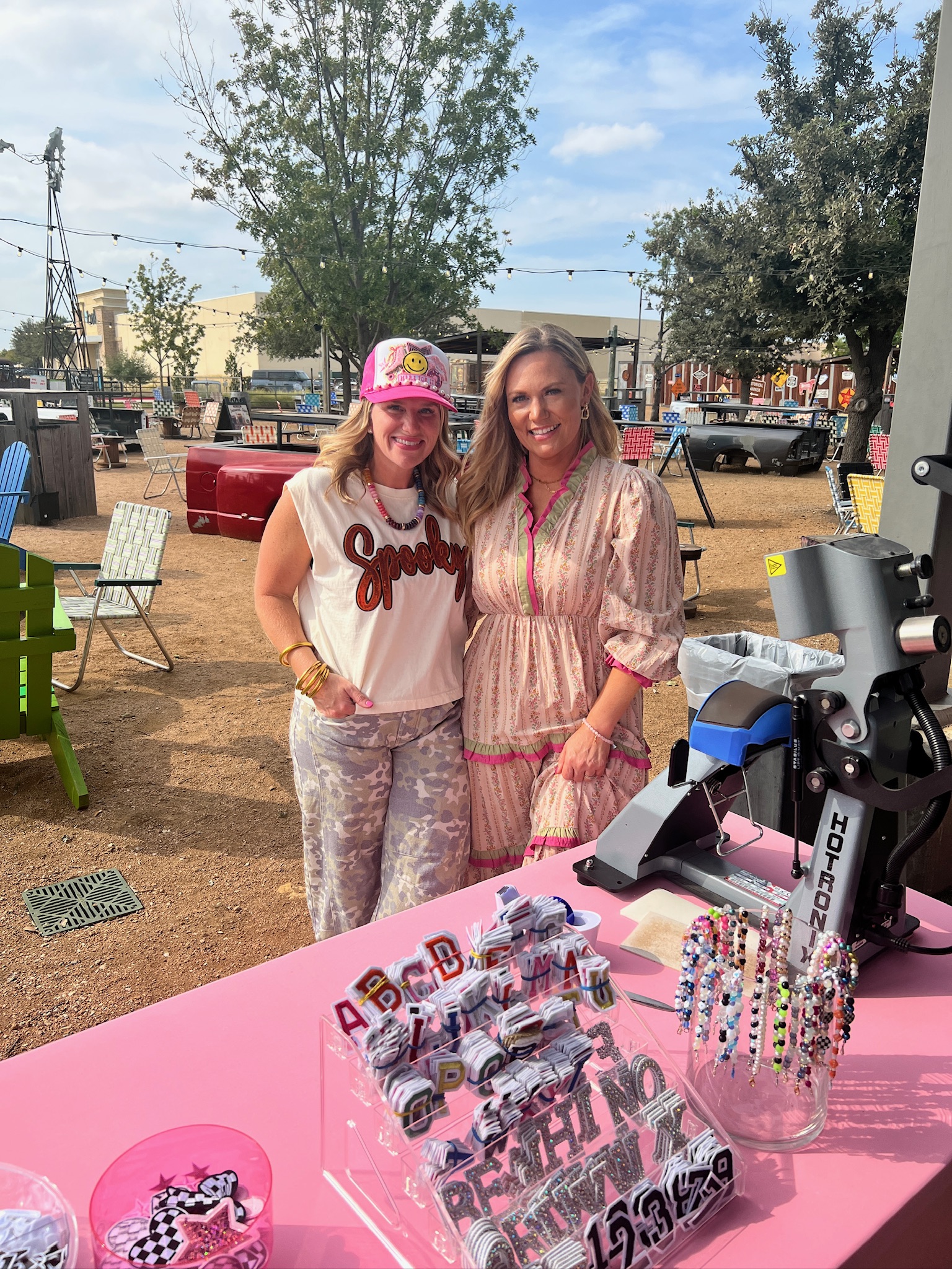 Audrie Dollins with her friend, owner of Southern Junkiez, at The Mom Game Live event at Truck Yard Fort Worth, standing by the custom hat bar supporting Susan G. Komen for breast cancer awareness.
