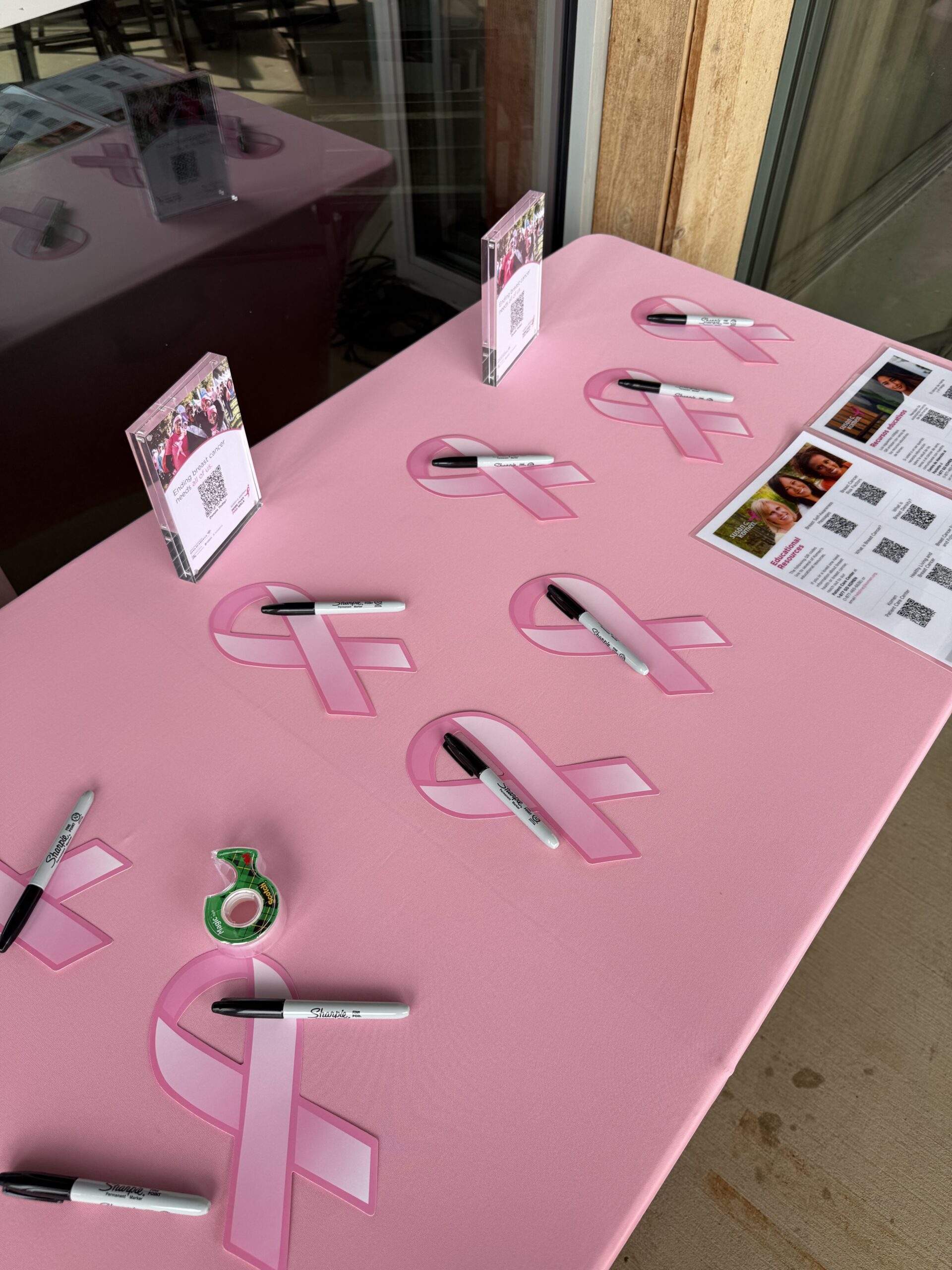 Pink ribbon table at The Mom Game Live event where guests wrote names to honor loved ones affected by breast cancer and placed ribbons on a window display supporting Susan G. Komen.