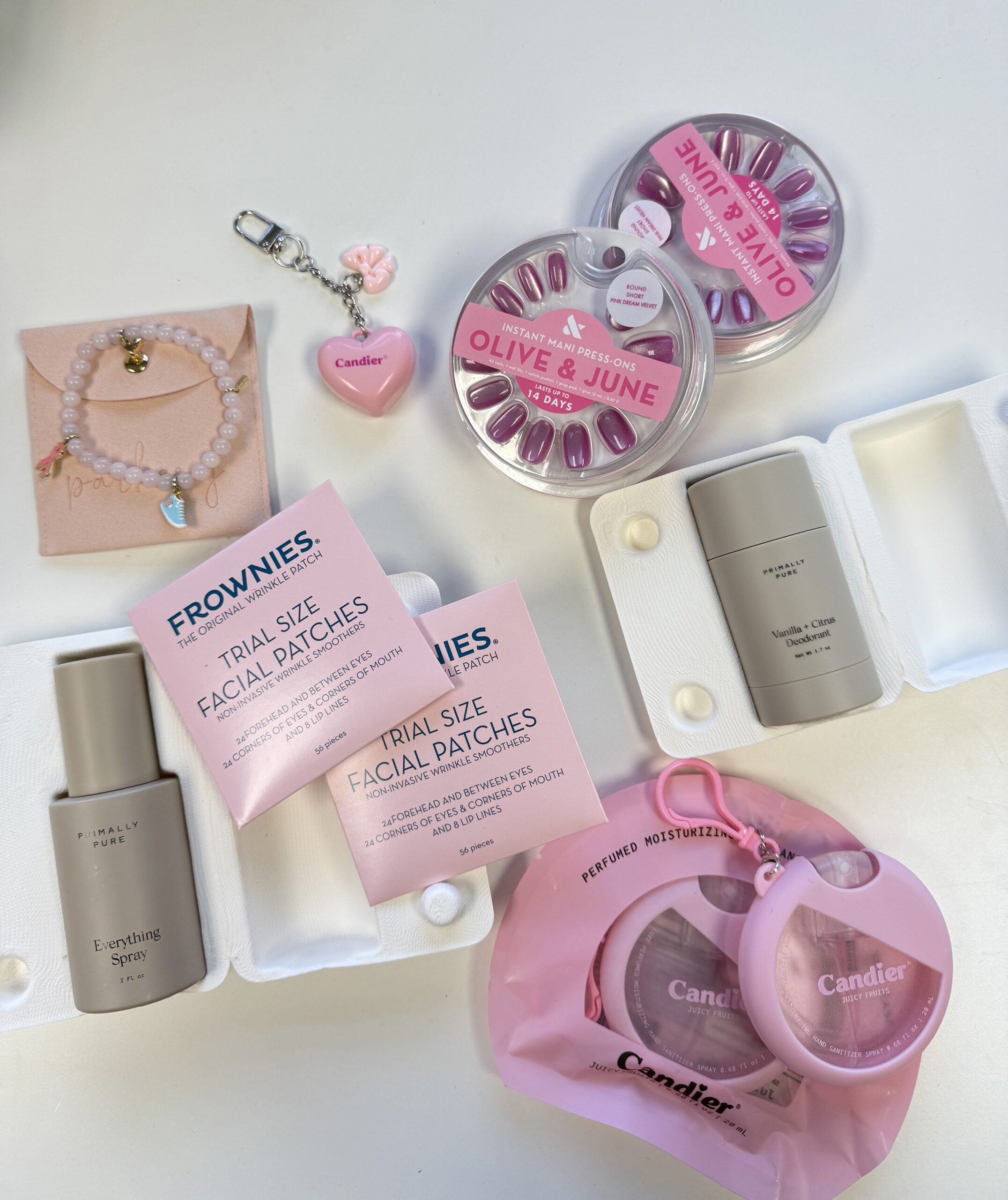 Flat lay of The Mom Game Live event swag bag items including Olive & June press-on nails, Parker J PJ bracelet, The Candier heart-shaped lip balm, Primally Pure Everything Spray and deodorant, Frownies facial patches, and Petal & Pup and Commense fashion pieces, created in partnership with Susan G. Komen at Truck Yard Fort Worth.