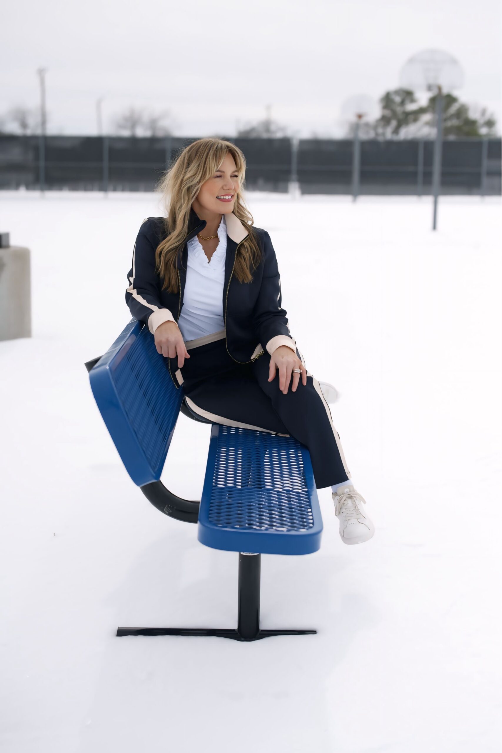 Audrie Dollins seated on a blue bench outdoors in a snowy setting, wearing a navy jacket, white blouse, and sneakers, discussing Facebook conversion strategy, buyer intent, and revenue focused social media performance.