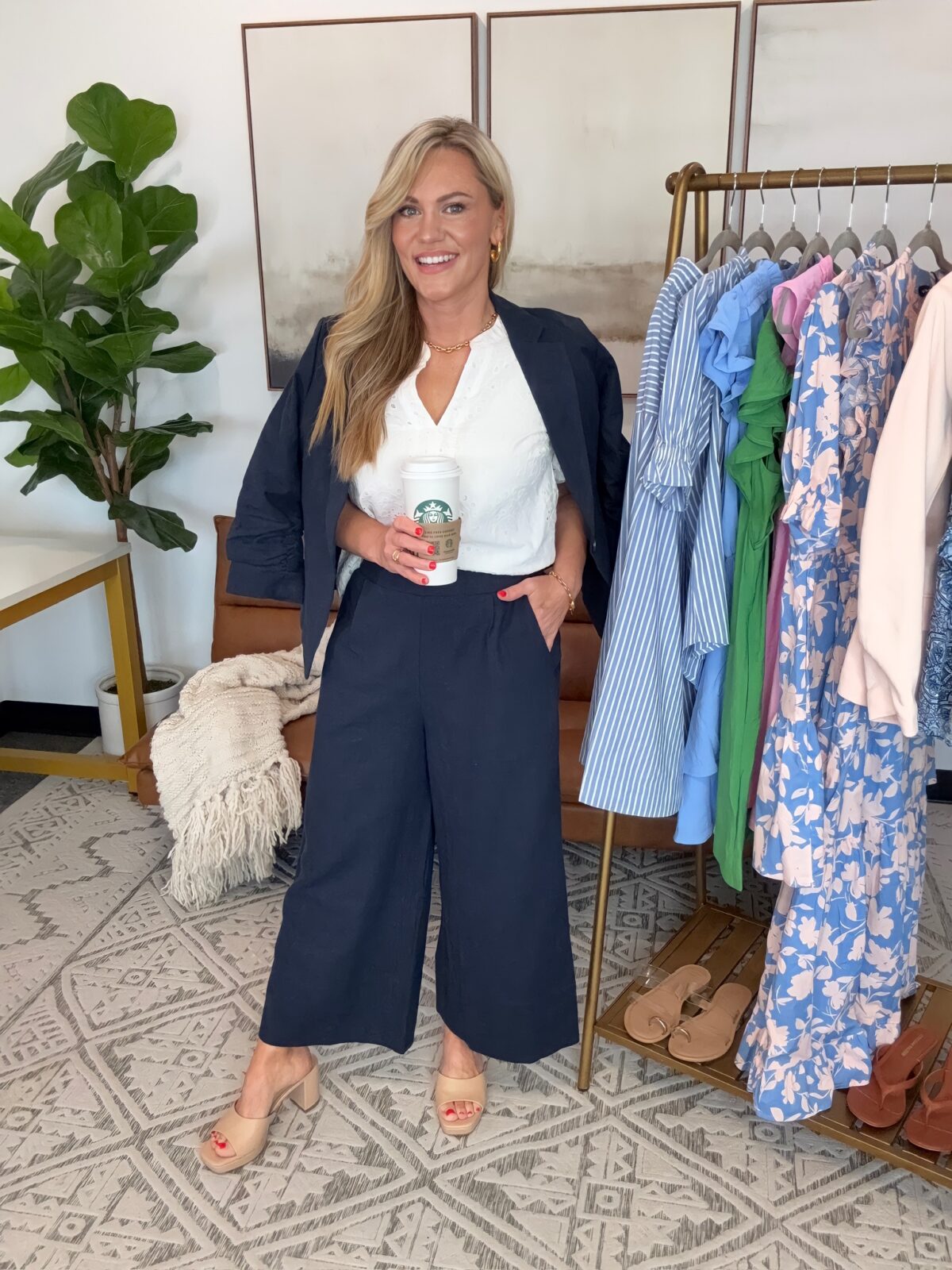 Audrie Dollins wearing a navy and white business casual outfit with a cropped blazer, white sleeveless top, wide leg culotte trousers, and nude heels in a boutique inspired office setting. Posing with coffee beside a rack of spring clothing and floral dresses.