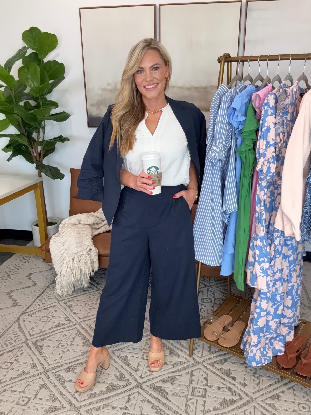 Audrie Dollins wearing a navy cropped blazer with matching navy wide leg culotte pants, a white textured sleeveless blouse, and nude block heel sandals while holding a Starbucks coffee. Standing beside a clothing rack filled with spring dresses and striped pieces in a bright styled office space.