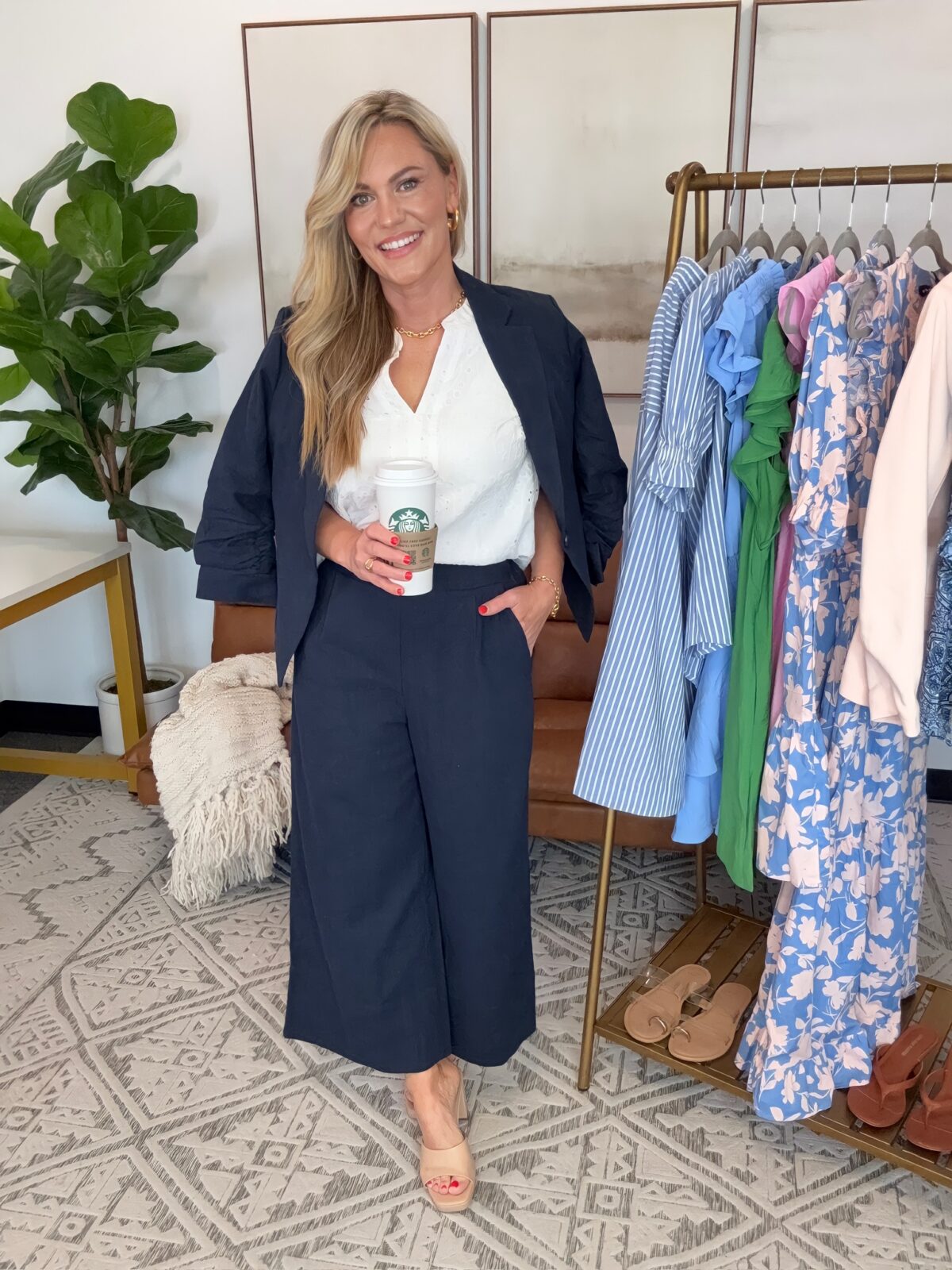 Audrie Dollins styled in a business casual spring outfit featuring a navy blazer, white sleeveless blouse, navy cropped wide leg pants, and neutral platform heels. Standing indoors next to a curated clothing rack with colorful dresses and spring fashion pieces.