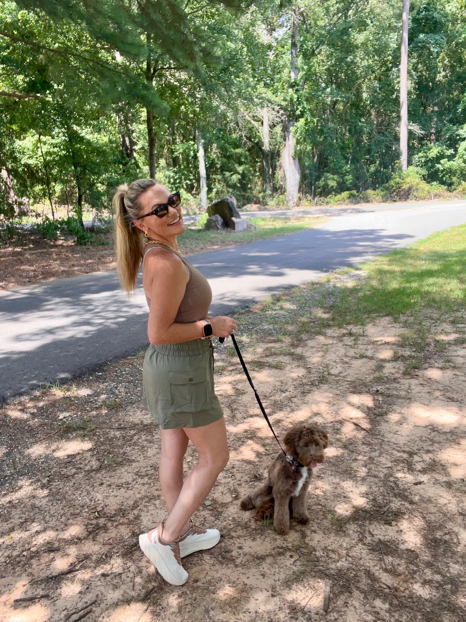 Woman wearing a brown fitted tank top and olive green cargo skort walking her dog in a wooded park, showcasing a comfortable summer activewear outfit with casual sneakers.