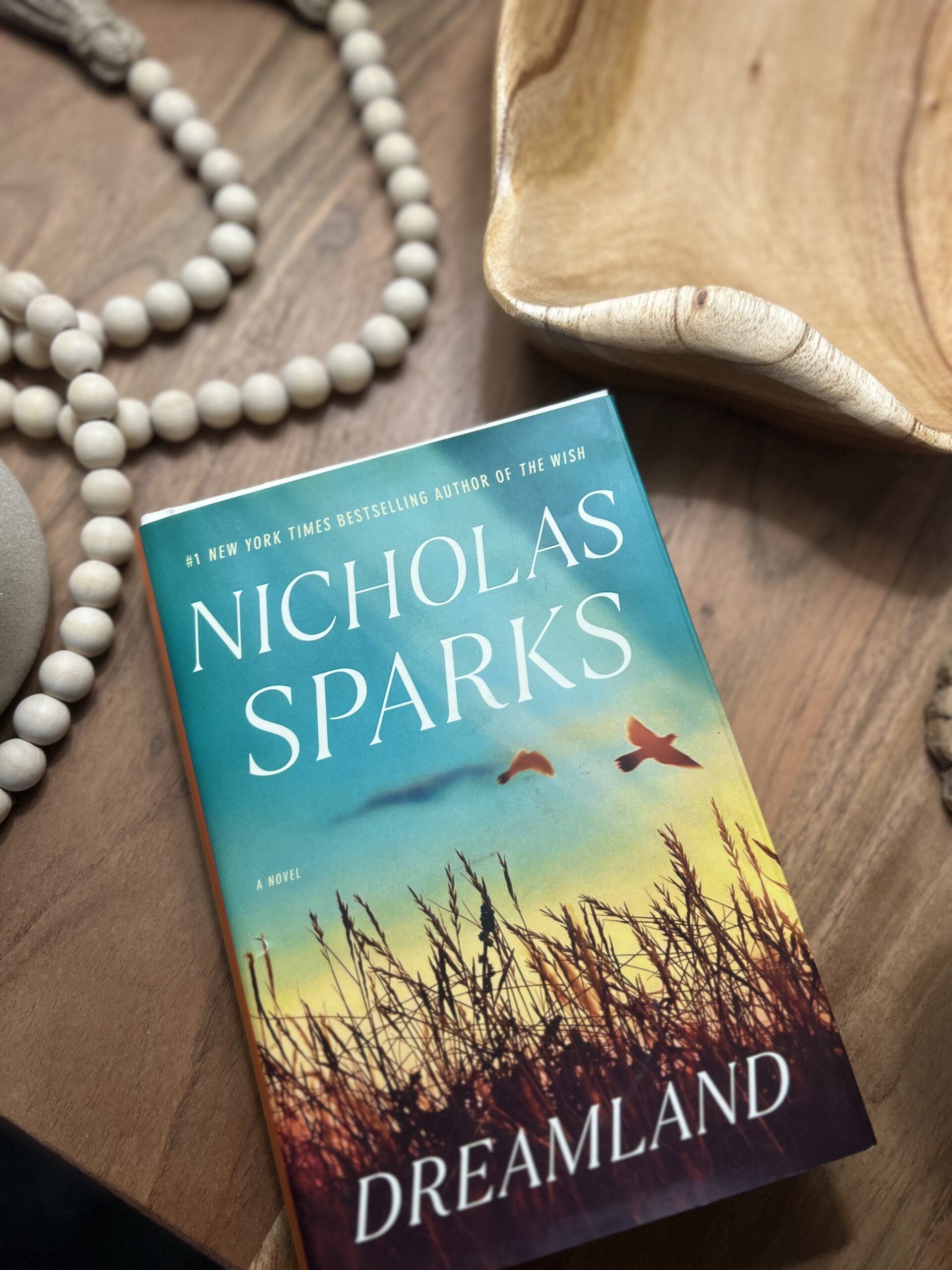 Nicholas Sparks' "Dreamland" arranged on a wooden table with decorative beads and a wooden tray, showcasing a bestselling romance novel for a summer reading list.