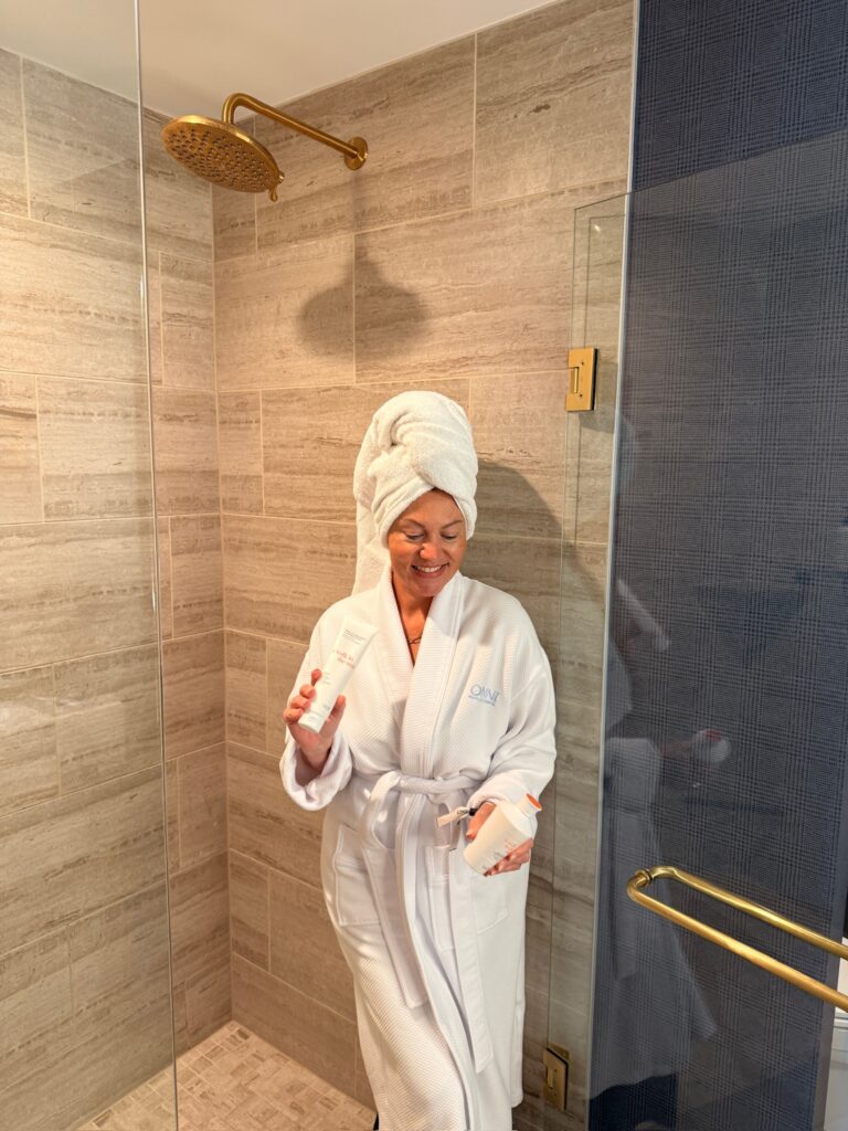 Audrie Dollins wearing a white robe and towel while holding Voir Hair Care Shampoo and Hair Mask at LTKCon 2026, highlighting a beauty brand collaboration through Parlay Collective.