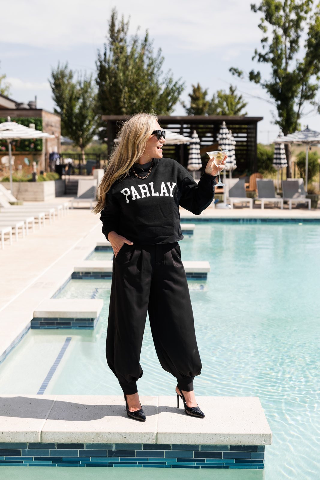 Audrie Dollins wearing a black PARLAY sweatshirt, black wide-leg pants, and pointed-toe heels, sitting on a white poolside lounge chair while holding a martini. Black-and-white striped umbrellas and rows of lounge chairs fill the background at a luxury resort.
